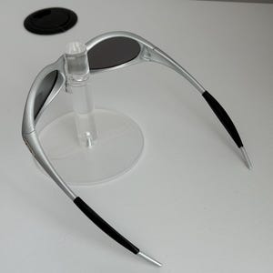 May include: Silver sunglasses with dark lenses. The sunglasses have a sleek design with black accents on the temples. The glasses are displayed on a clear plastic stand.