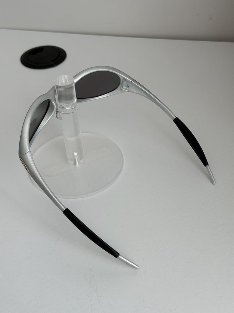 May include: Silver sunglasses with dark lenses. The sunglasses have a sleek design with black accents on the temples. The glasses are displayed on a clear plastic stand.