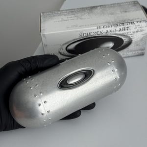 May include: Silver-coloured eyewear case with the Oakley logo. The case has a cylindrical shape with rivets and a black logo. A white box with text is in the background.