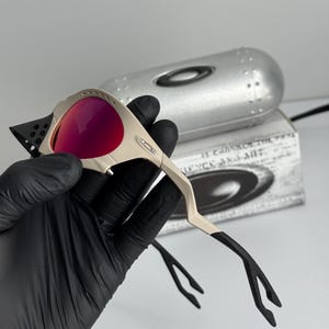 Może przedstawiać: Okulary przeciwsłoneczne z beżową oprawką i czerwonymi soczewkami. Okulary mają unikalny, kanciasty design z czarnymi akcentami na zausznikach. Srebrne etui i pudełko z tekstem znajdują się w tle.