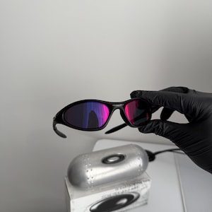 May include: Black sunglasses with a curved frame and reflective pink and purple lenses. The sunglasses are held by a gloved hand. A silver case and a white box with a black logo are below the sunglasses.
