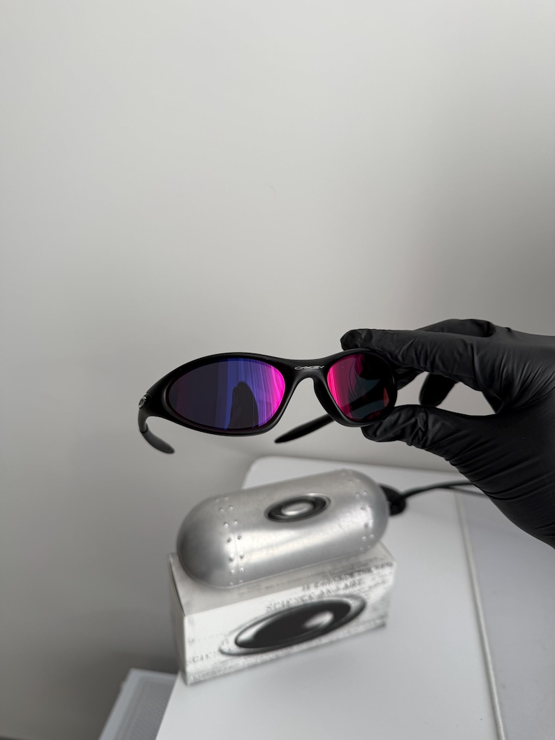 May include: Black sunglasses with a curved frame and reflective pink and purple lenses. The sunglasses are held by a gloved hand. A silver case and a white box with a black logo are below the sunglasses.