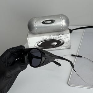 K&ouml;nnte beinhalten: Schwarze Sonnenbrille mit Wrap-around-Design, dunklen Gl&auml;sern und einem silbernen Etui. Das Etui und die Schachtel tragen das Markenlogo. Auf der Schachtel steht "SCIENCE AND ART".