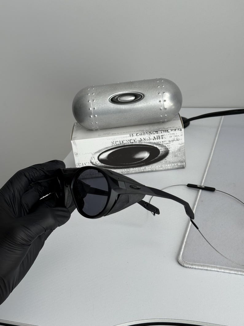 K&ouml;nnte beinhalten: Schwarze Sonnenbrille mit Wrap-around-Design, dunklen Gl&auml;sern und einem silbernen Etui. Das Etui und die Schachtel tragen das Markenlogo. Auf der Schachtel steht "SCIENCE AND ART".