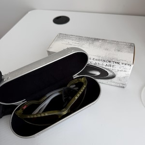 May include: Open silver sunglasses case with black interior, holding olive green and white sunglasses with dark lenses. A white box with text is behind the case. The sunglasses are a sporty style.