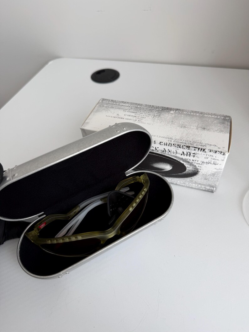 May include: Open silver sunglasses case with black interior, holding olive green and white sunglasses with dark lenses. A white box with text is behind the case. The sunglasses are a sporty style.