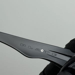 May include: Close-up of a black eyeglass arm with etched text and symbols. The arm is held by a gloved hand against a light gray background. The text is small and difficult to read.