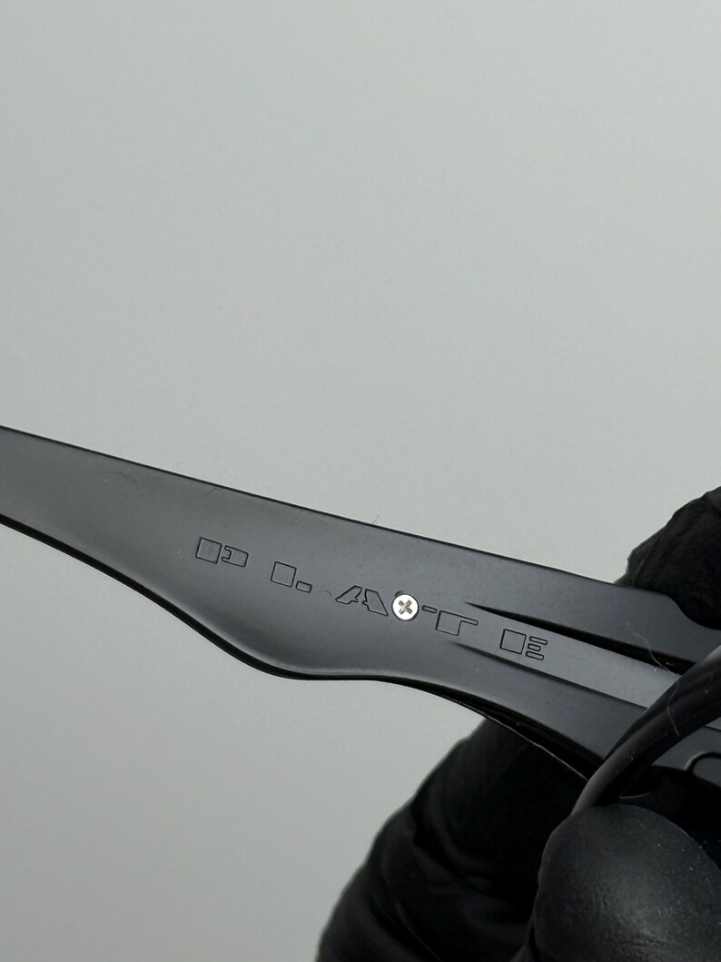 May include: Close-up of a black eyeglass arm with etched text and symbols. The arm is held by a gloved hand against a light gray background. The text is small and difficult to read.