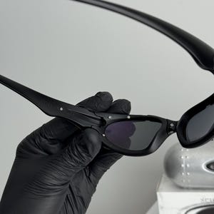 May include: Black sunglasses with a curved frame and dark lenses. The sunglasses are held by a gloved hand, showcasing the sleek design and details. The lenses appear to be tinted, offering a stylish look.
