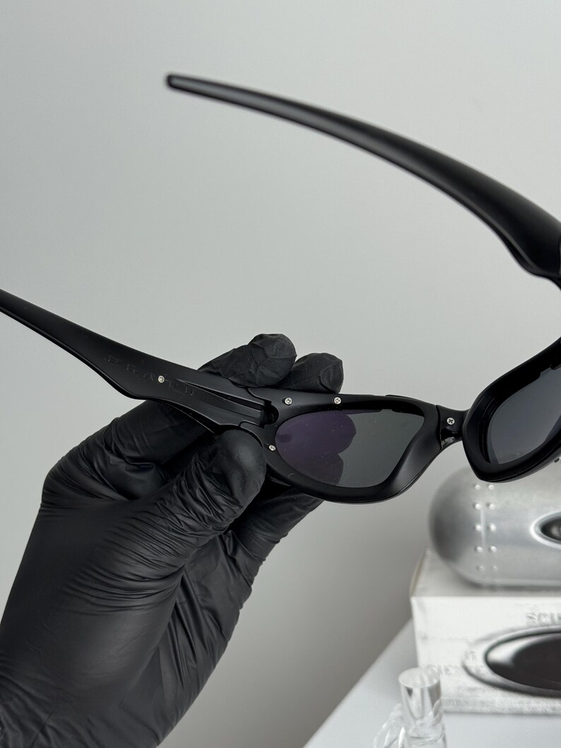 May include: Black sunglasses with a curved frame and dark lenses. The sunglasses are held by a gloved hand, showcasing the sleek design and details. The lenses appear to be tinted, offering a stylish look.