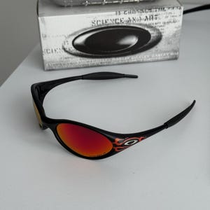 May include: Black sunglasses with red lenses and flame design accents. The sunglasses are set against a white background, with a box and a metallic object in the background. The text on the box reads "SCIENCE AND ART".