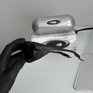 K&ouml;nnte beinhalten: Schwarze Sonnenbrille und ein silbernes Etui. Die Sonnenbrille hat ein modernes Design mit dunklen Gl&auml;sern und einem schwarzen Rahmen. Das Etui ist silberfarben mit einem Nietendesign und dem Oakley-Logo. Auf der Box steht "SCIENCE AND ART".