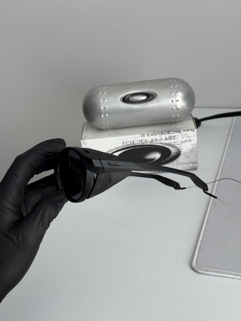 K&ouml;nnte beinhalten: Schwarze Sonnenbrille und ein silbernes Etui. Die Sonnenbrille hat ein modernes Design mit dunklen Gl&auml;sern und einem schwarzen Rahmen. Das Etui ist silberfarben mit einem Nietendesign und dem Oakley-Logo. Auf der Box steht "SCIENCE AND ART".