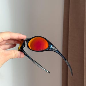 Puede incluir: Gafas de sol deportivas con montura negra y azul y lentes rojas vibrantes. El dise&ntilde;o elegante presenta una forma curvada y una fina patilla plateada. Las lentes ofrecen un contraste llamativo con la montura oscura, creando un look moderno.