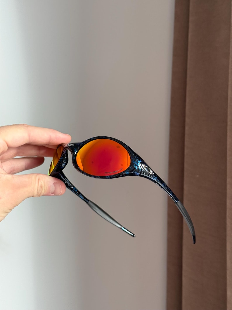 Puede incluir: Gafas de sol deportivas con montura negra y azul y lentes rojas vibrantes. El dise&ntilde;o elegante presenta una forma curvada y una fina patilla plateada. Las lentes ofrecen un contraste llamativo con la montura oscura, creando un look moderno.