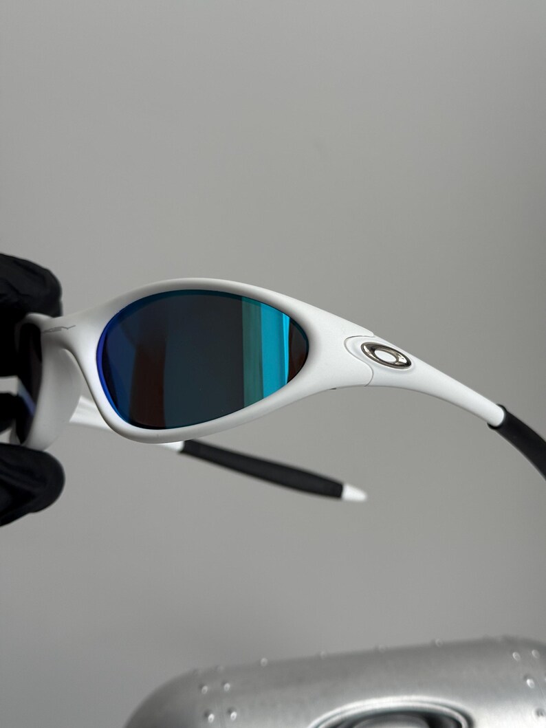 May include: White sunglasses with blue mirrored lenses. The sunglasses have a sleek, modern design with a white frame and black arms. The lenses reflect shades of blue and green.