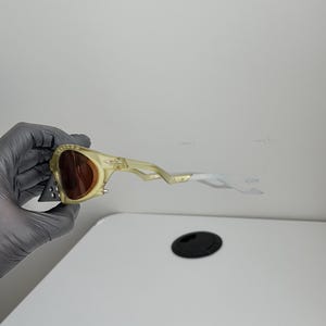 May include: Yellow-tinted sunglasses with brown lenses. The sunglasses feature an unusual, angular design with a clear, white arm and a small, triangular detail on the frame. The lenses are a warm brown colour.