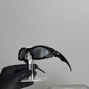 May include: Black sunglasses with a curved design and reflective lenses. The sunglasses are held on a clear stand, with a silver case in the background. The sunglasses have a sleek, modern aesthetic.