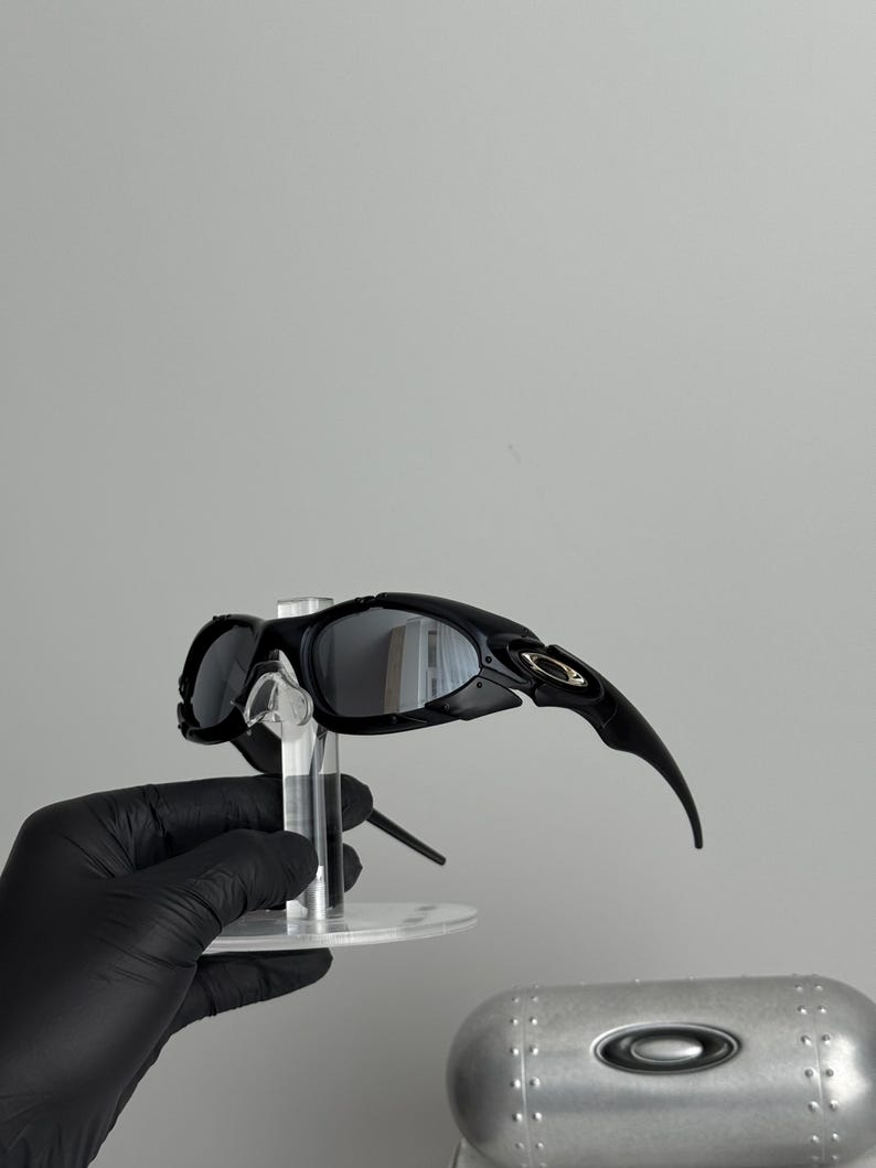 May include: Black sunglasses with a curved design and reflective lenses. The sunglasses are held on a clear stand, with a silver case in the background. The sunglasses have a sleek, modern aesthetic.