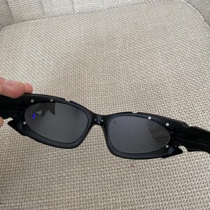 May include: Black sunglasses with a distinctive, angular design. The frames are matte black with small silver accents. The lenses are dark, offering a sleek, modern appearance. These sunglasses are a fashionable accessory.