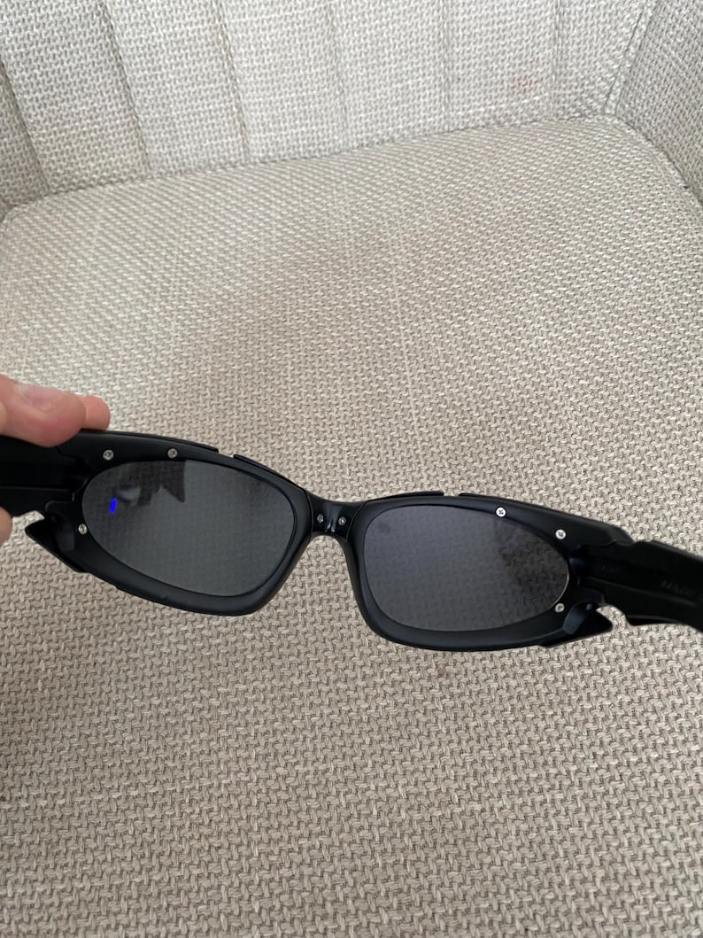 May include: Black sunglasses with a distinctive, angular design. The frames are matte black with small silver accents. The lenses are dark, offering a sleek, modern appearance. These sunglasses are a fashionable accessory.