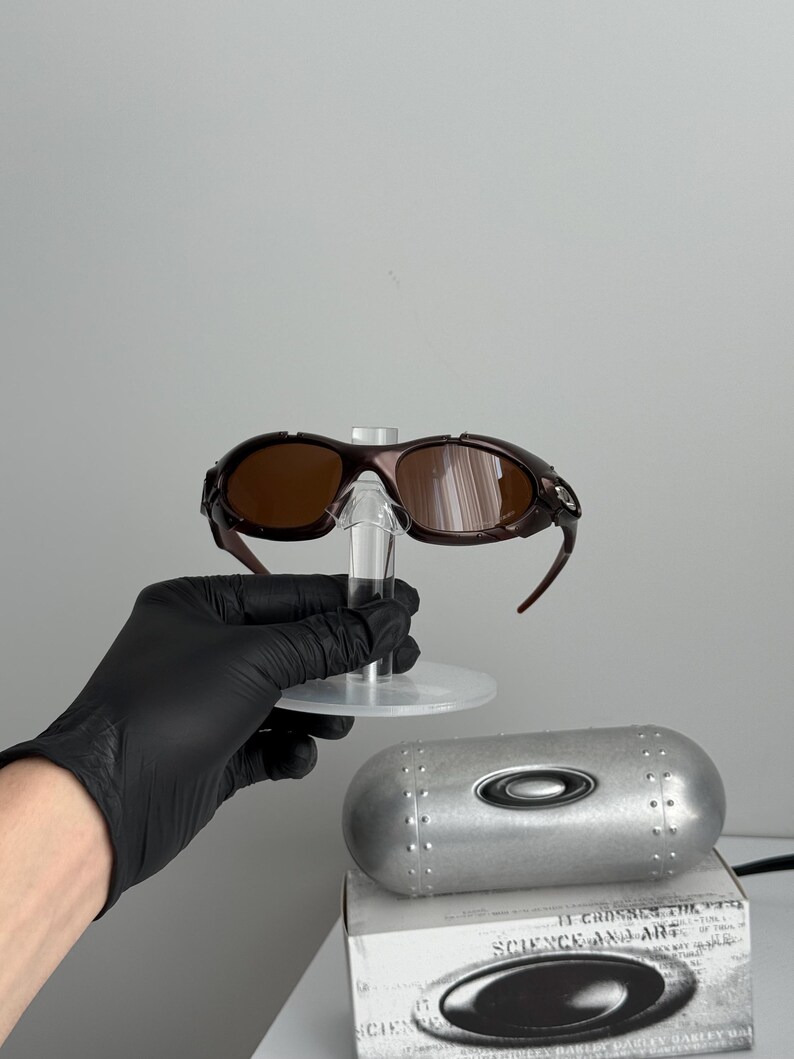 May include: Brown-tinted sunglasses with a dark brown frame, displayed on a clear stand. A silver case and a white box with the text "SCIENCE AND ART" are also visible. The sunglasses have a sporty design.