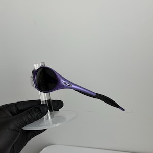 May include: Purple and black sunglasses with a sleek, modern design. The sunglasses have a curved frame with a gradient from purple to black. The lenses are dark, and the arms are thin and streamlined.