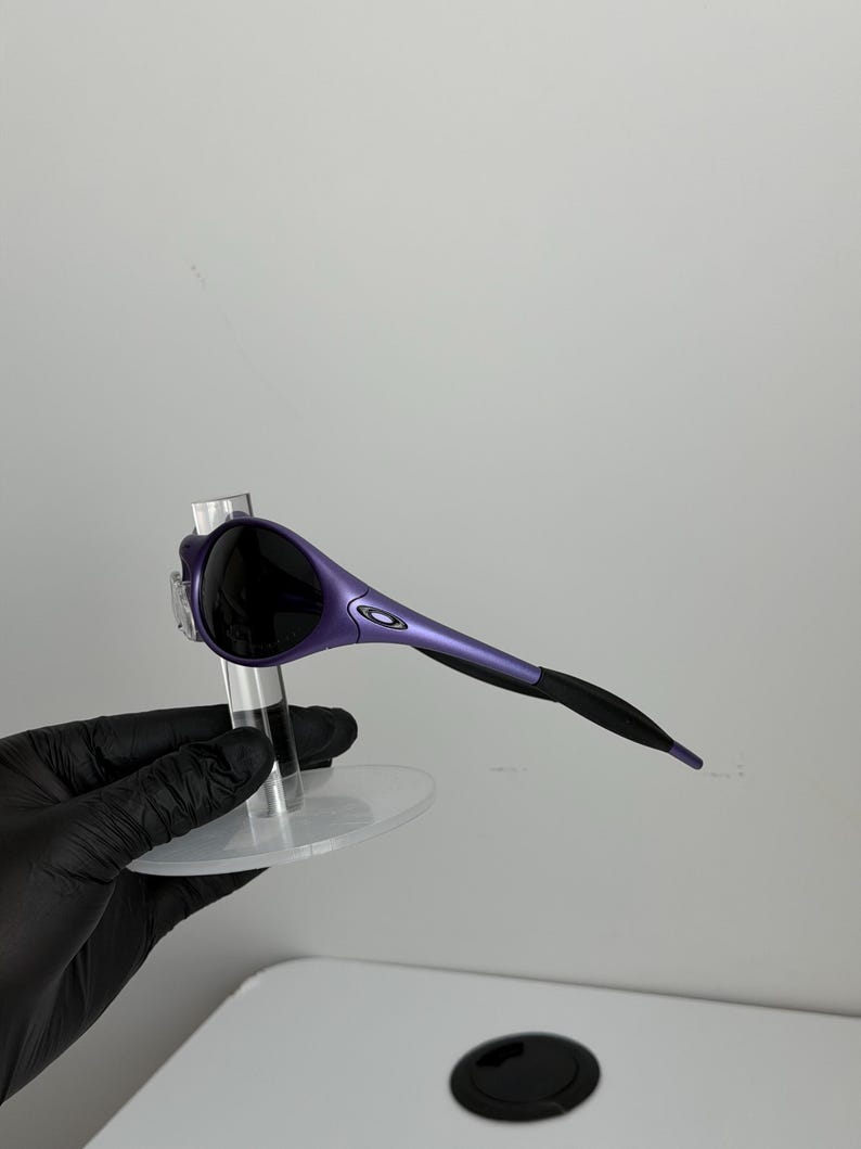 May include: Purple and black sunglasses with a sleek, modern design. The sunglasses have a curved frame with a gradient from purple to black. The lenses are dark, and the arms are thin and streamlined.