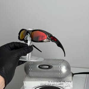 May include: Black and silver sunglasses with red and yellow lenses. The sunglasses are displayed on a clear stand, with a silver case and a box with the text "SCIENCE AND ART" visible in the background.