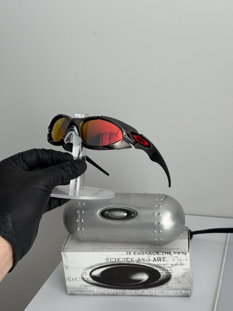 May include: Black and silver sunglasses with red and yellow lenses. The sunglasses are displayed on a clear stand, with a silver case and a box with the text "SCIENCE AND ART" visible in the background.