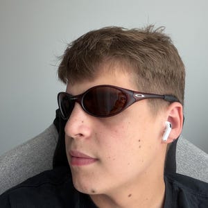 May include: Dark brown oval sunglasses with a black frame. The sunglasses have a reflective lens. A pair of white wireless earbuds are in the ear. The person is wearing a black shirt.
