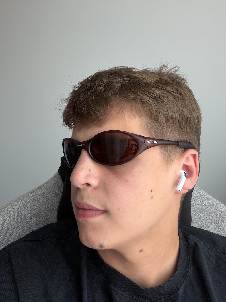 May include: Dark brown oval sunglasses with a black frame. The sunglasses have a reflective lens. A pair of white wireless earbuds are in the ear. The person is wearing a black shirt.