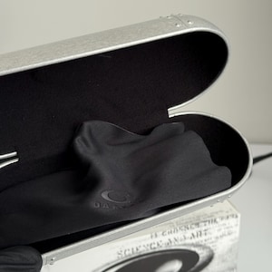 May include: Open silver-colored glasses case with a black interior and a black microfiber cloth. The case is oval-shaped with a riveted design. The cloth has the Oakley logo. A white box with the words "SCIENCE AND ART" is visible.