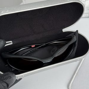 Puede incluir: Gafas de sol Oakley negras con lentes oscuras dentro de un estuche plateado abierto con interior negro. El estuche muestra las gafas de sol y un pa&ntilde;o de limpieza. Las gafas tienen una peque&ntilde;a marca "USA".