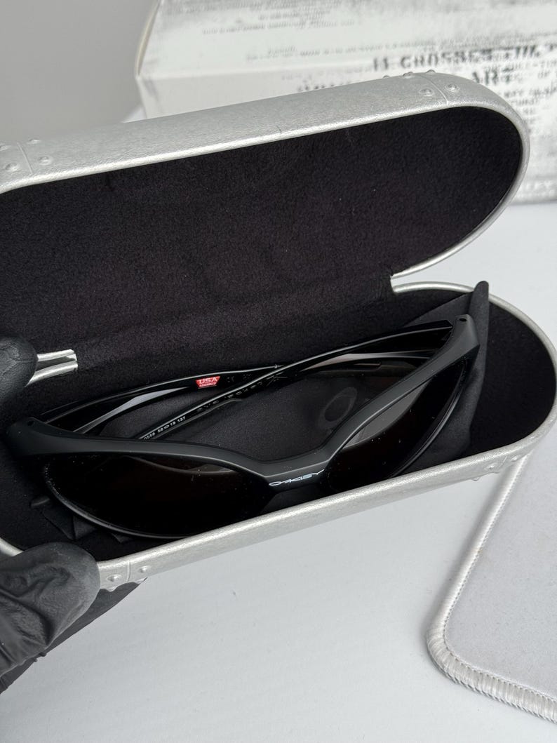 Puede incluir: Gafas de sol Oakley negras con lentes oscuras dentro de un estuche plateado abierto con interior negro. El estuche muestra las gafas de sol y un pa&ntilde;o de limpieza. Las gafas tienen una peque&ntilde;a marca "USA".