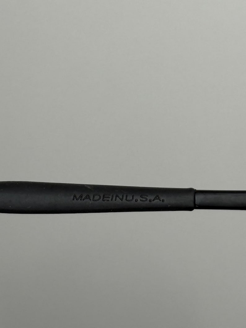 K&ouml;nnte beinhalten: Nahaufnahme eines schwarzen Brillenb&uuml;gels mit dem wei&szlig;en Aufdruck "MADE IN U.S.A.". Der B&uuml;gel besteht aus mattem Material und vor grauem Hintergrund.