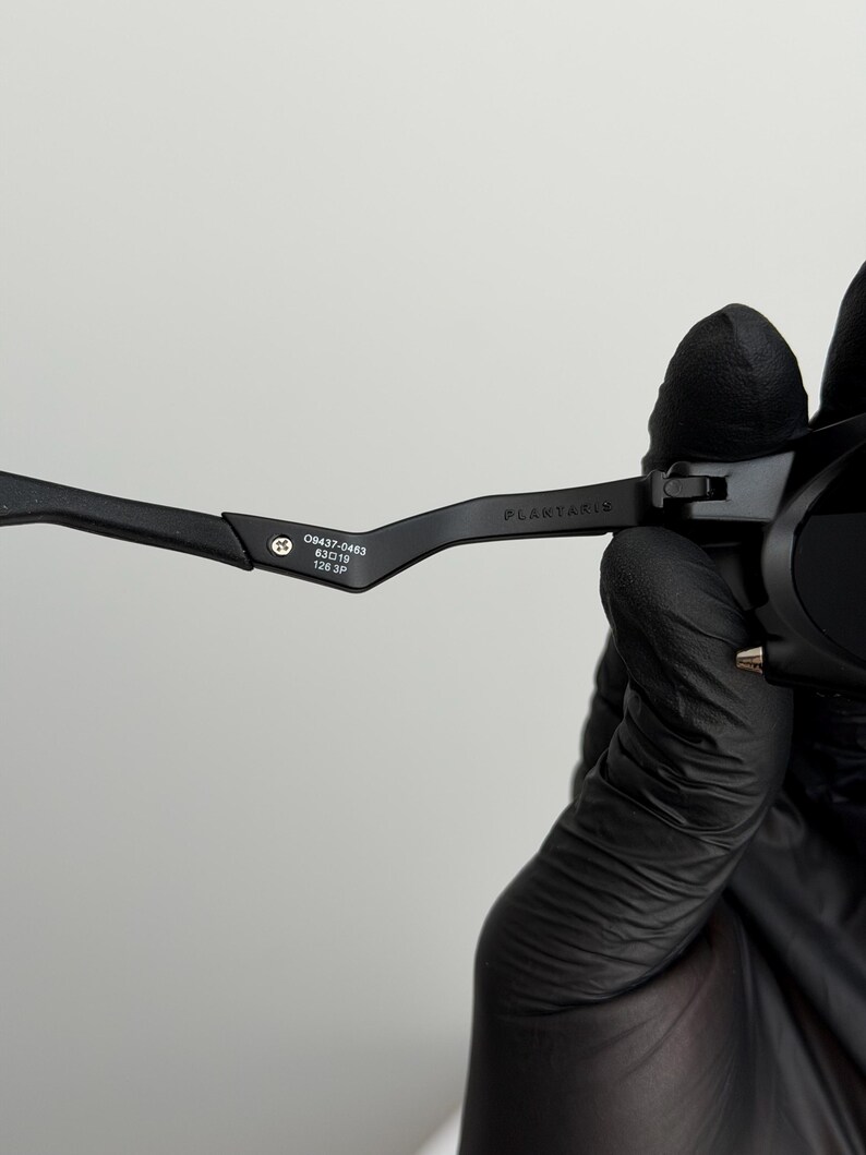 May include: Close-up of black sunglasses with the text "PLANTARIS" on the temple. The glasses have the numbers "09437-0463 63▢19 126 3P" printed on the arm. The glasses are held by a person wearing a black glove.