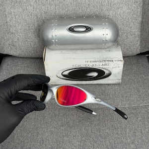 May include: Silver-framed sunglasses with vibrant red-orange lenses. The glasses are displayed with a silver case and a box that reads "SCIENCE AND ART". The sunglasses have black accents on the arms.