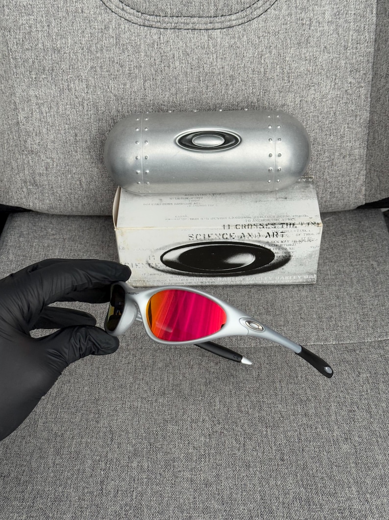May include: Silver-framed sunglasses with vibrant red-orange lenses. The glasses are displayed with a silver case and a box that reads "SCIENCE AND ART". The sunglasses have black accents on the arms.
