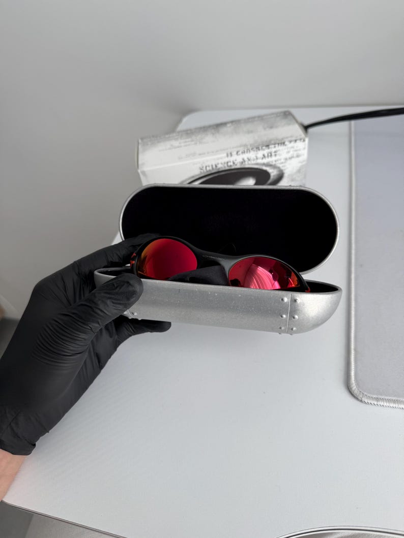 May include: Red-lensed sunglasses in a silver case. The sunglasses have a black frame and are in an open, silver case with a black interior. The case is held by a gloved hand. A white package with text is in the background.