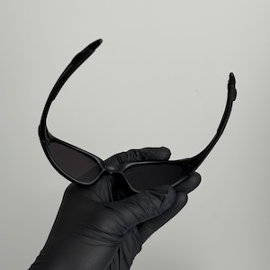 K&ouml;nnte beinhalten: Schwarze Sonnenbrille mit einem schlanken, modernen Design. Die Sonnenbrille hat einen gebogenen Rahmen und dunkle Gl&auml;ser. Das Bild zeigt die Sonnenbrille, die von einer behandschuhten Hand vor einem schlichten Hintergrund gehalten wird. Ein stilvolles Accessoire.
