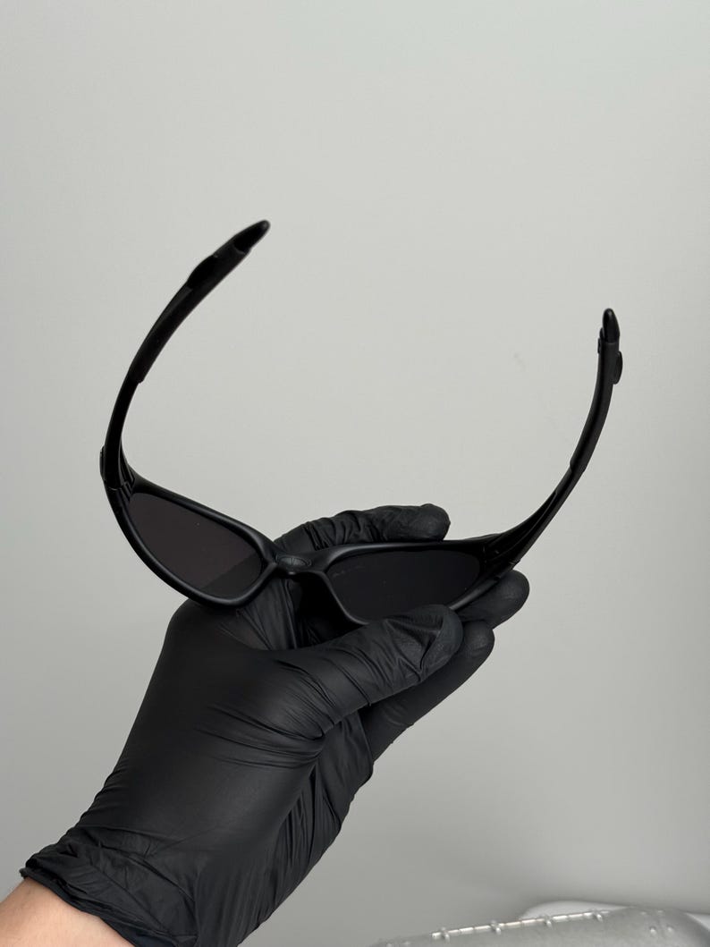 K&ouml;nnte beinhalten: Schwarze Sonnenbrille mit einem schlanken, modernen Design. Die Sonnenbrille hat einen gebogenen Rahmen und dunkle Gl&auml;ser. Das Bild zeigt die Sonnenbrille, die von einer behandschuhten Hand vor einem schlichten Hintergrund gehalten wird. Ein stilvolles Accessoire.
