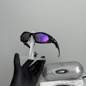 May include: Black Oakley sunglasses with purple mirrored lenses are displayed on a clear stand. The sunglasses are paired with a silver case and a box with the text "SCIENCE AND ART".