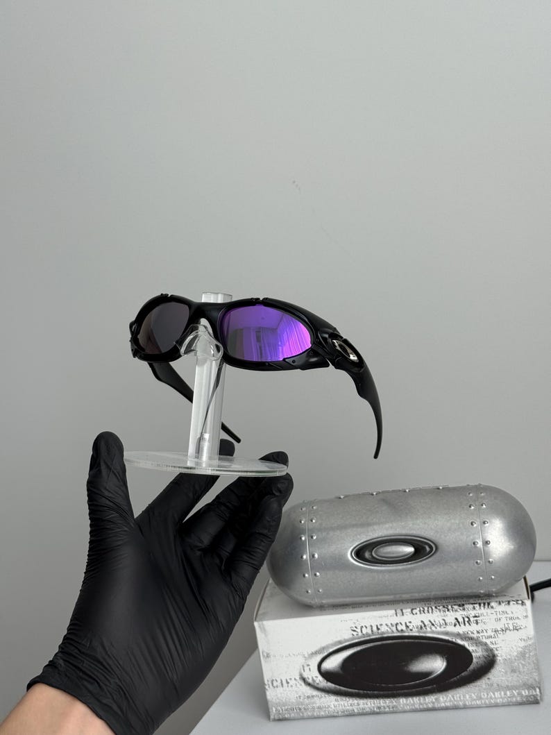 May include: Black Oakley sunglasses with purple mirrored lenses are displayed on a clear stand. The sunglasses are paired with a silver case and a box with the text "SCIENCE AND ART".