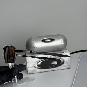 May include: A pair of Oakley sunglasses with a reflective lens, displayed with a silver case and a box. The sunglasses have a black frame and a silver accent. The case is silver with a black Oakley logo.