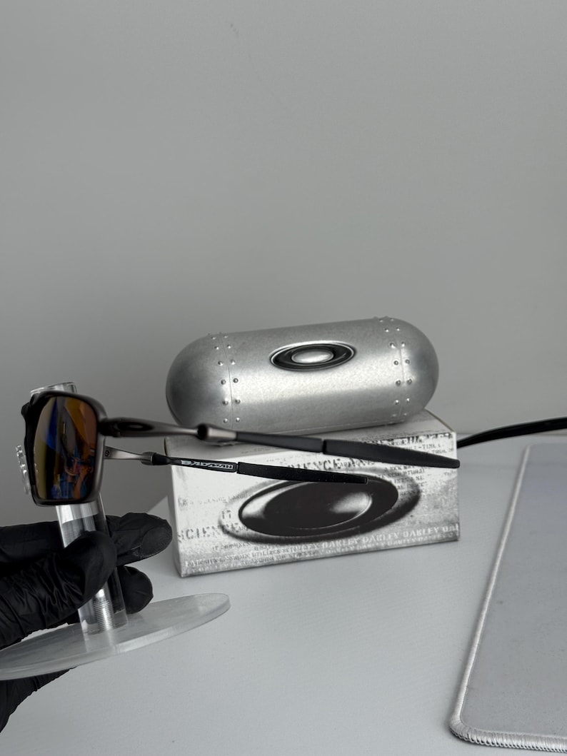 May include: A pair of Oakley sunglasses with a reflective lens, displayed with a silver case and a box. The sunglasses have a black frame and a silver accent. The case is silver with a black Oakley logo.
