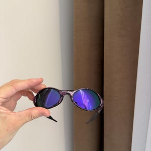 May include: Oval-shaped sunglasses with a black frame and purple and pink floral pattern. The lenses have a reflective blue hue. The sunglasses are held up by a hand.