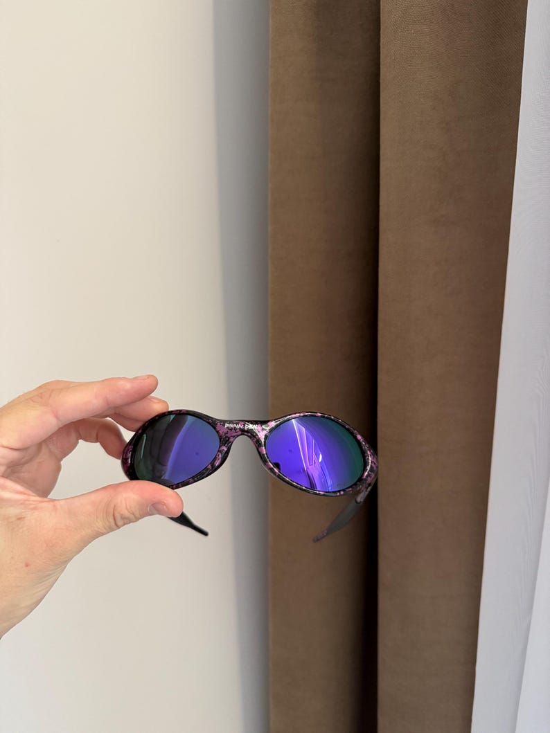 May include: Oval-shaped sunglasses with a black frame and purple and pink floral pattern. The lenses have a reflective blue hue. The sunglasses are held up by a hand.