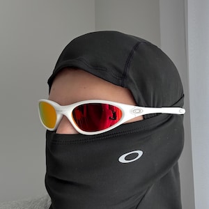 May include: White sunglasses with red mirrored lenses. The sunglasses are worn with a black balaclava. The Oakley logo is visible on the balaclava and the sunglasses' temples. The lenses reflect a warm, orange hue.