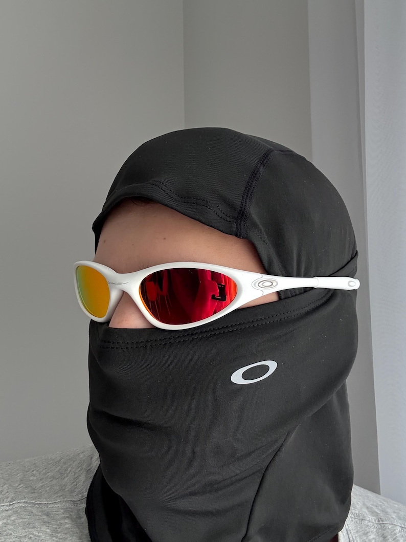 May include: White sunglasses with red mirrored lenses. The sunglasses are worn with a black balaclava. The Oakley logo is visible on the balaclava and the sunglasses' temples. The lenses reflect a warm, orange hue.